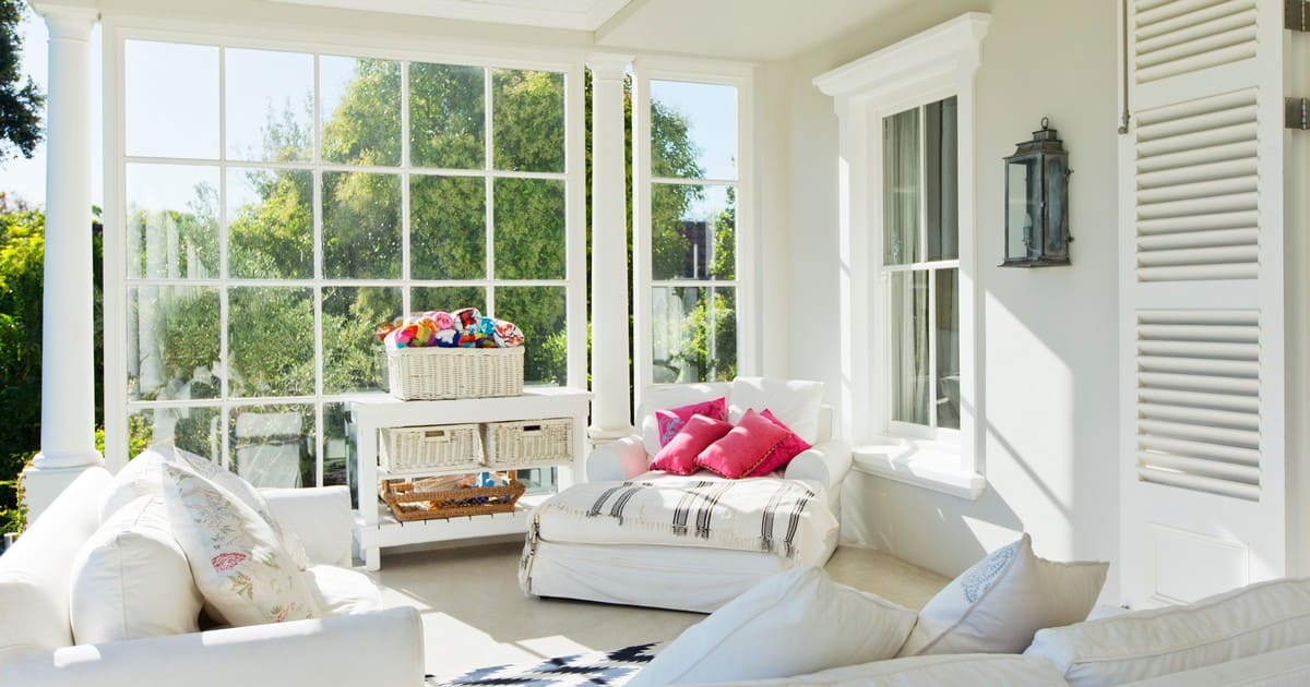 Bright sunroom with large floor-to-ceiling windows, white slipcovered seating, and pink accent pillows, illustrating a beautiful space for an ADU vs Home Addition Bay Area project.