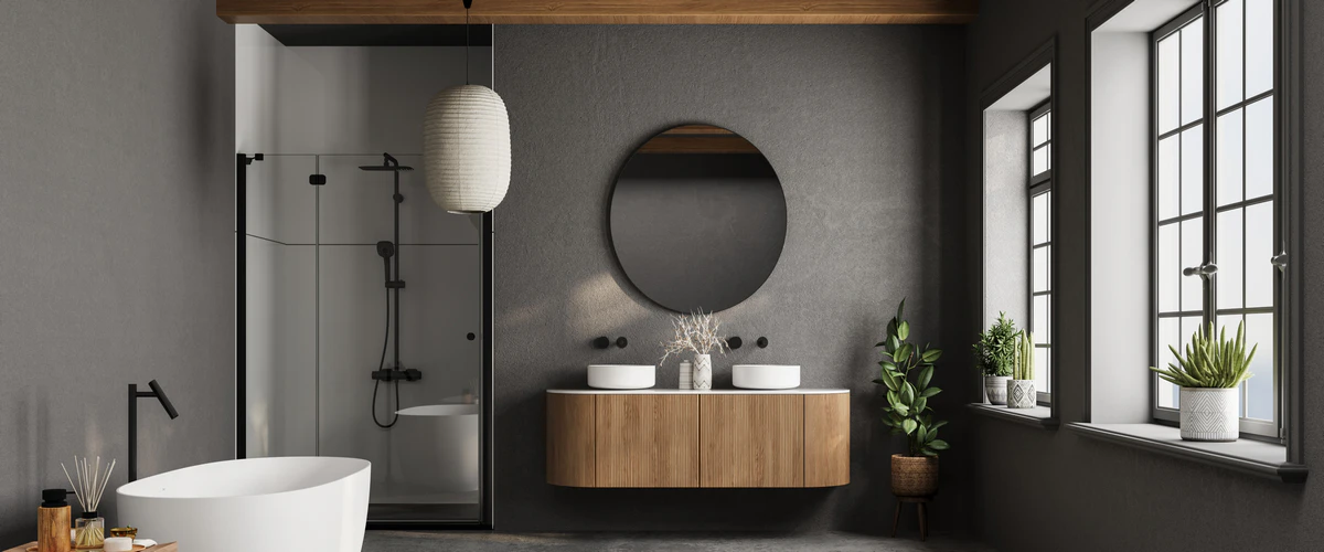 Contemporary dark gray bathroom featuring a slatted wood floating vanity with double vessel sinks, a white freestanding soaking tub, and a large round mirror by a bright window.
