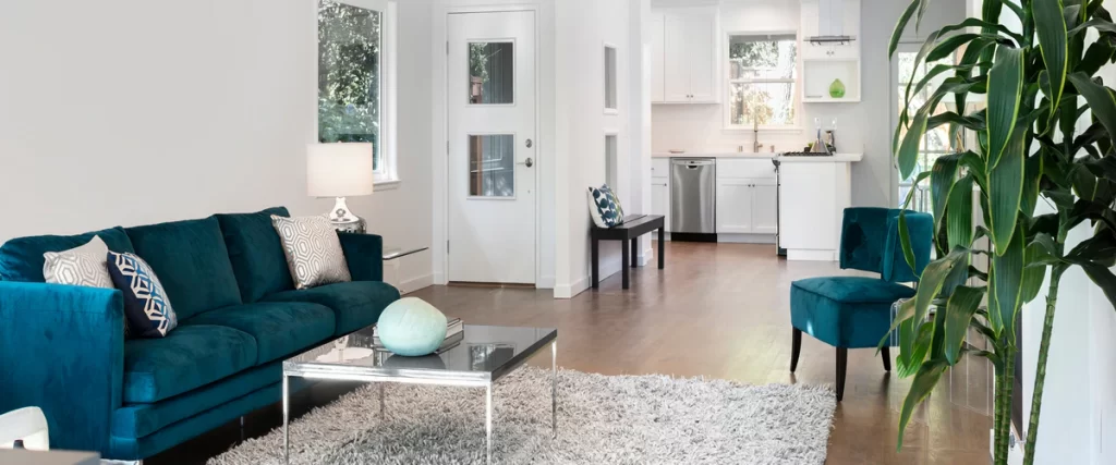 Modern open concept living room featuring a teal velvet sofa, matching accent chair, and glass coffee table as part of a bright new home addition leading into a white kitchen.