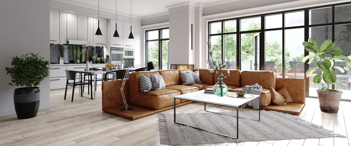 A spacious open-concept living room featuring a large tan leather sectional sofa, marble coffee table, and seamless transition into a modern white kitchen with large windows.