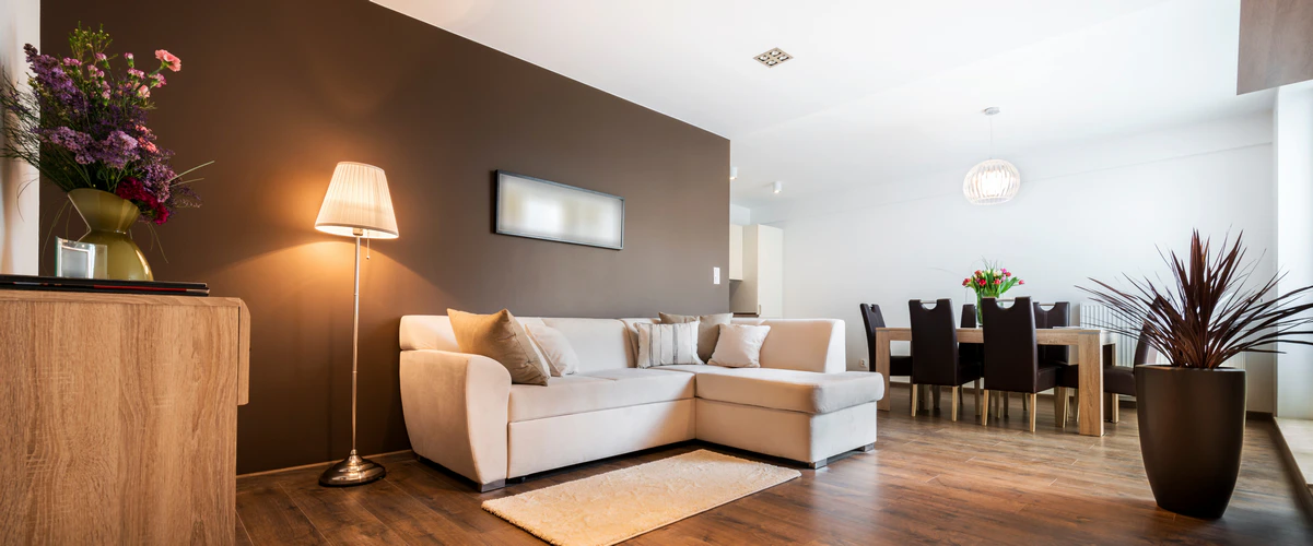 An open concept living room featuring a white sectional sofa against a dark brown accent wall, a floor lamp, hardwood floors, and a view into the adjacent dining room area.