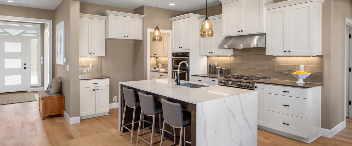Spacious transitional kitchen featuring white shaker cabinets, a large marble-look island with gray bar stools, and stainless steel appliances against warm beige walls.